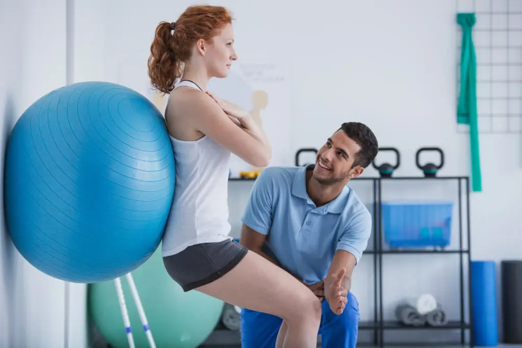 Physical Therapist working with a yound woman who is using an exersize ball against the wall for support.